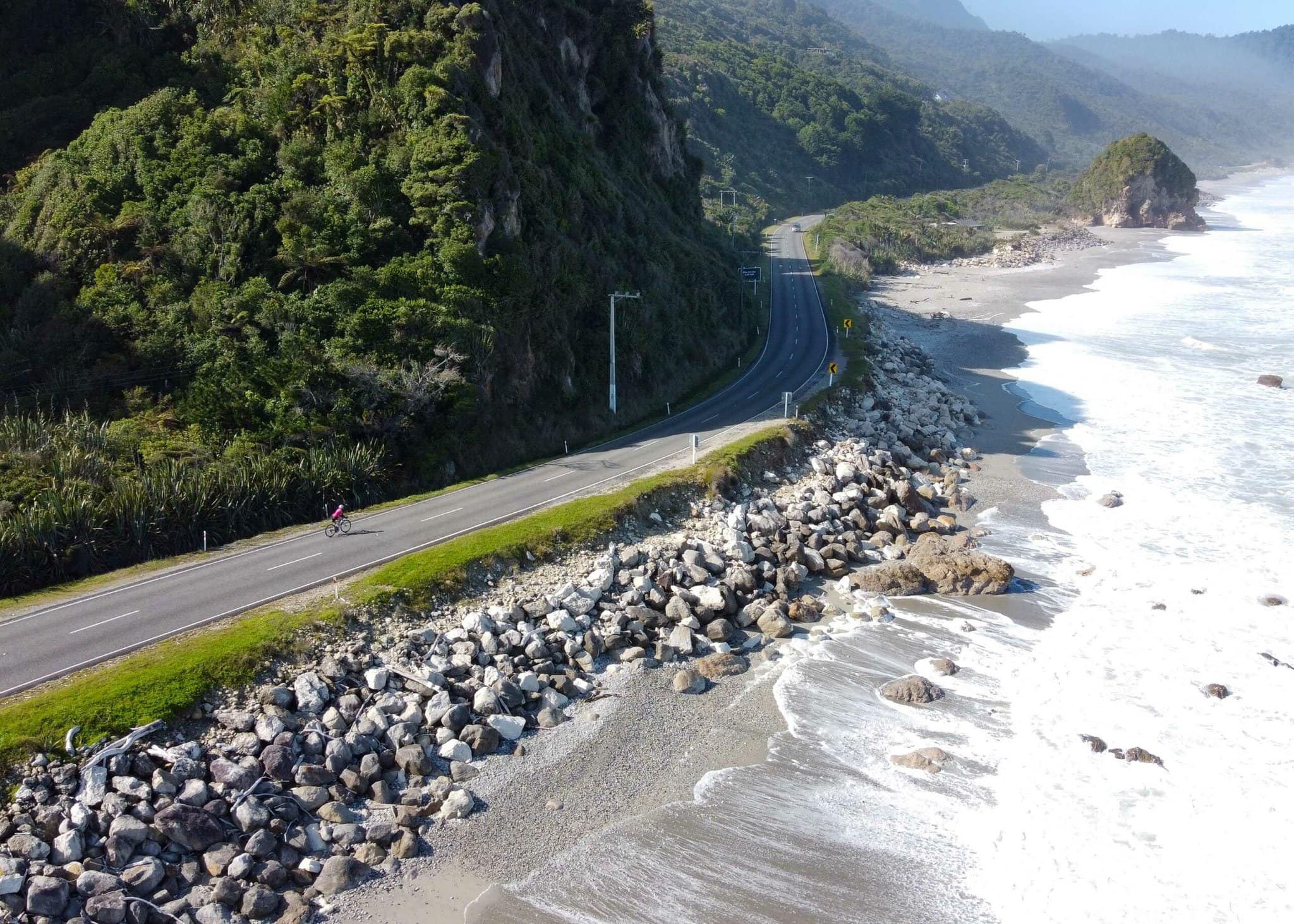 A cyclist rides along a road built along a rocky beach in New Zealand. The tall green hills come down to the edge of the road.