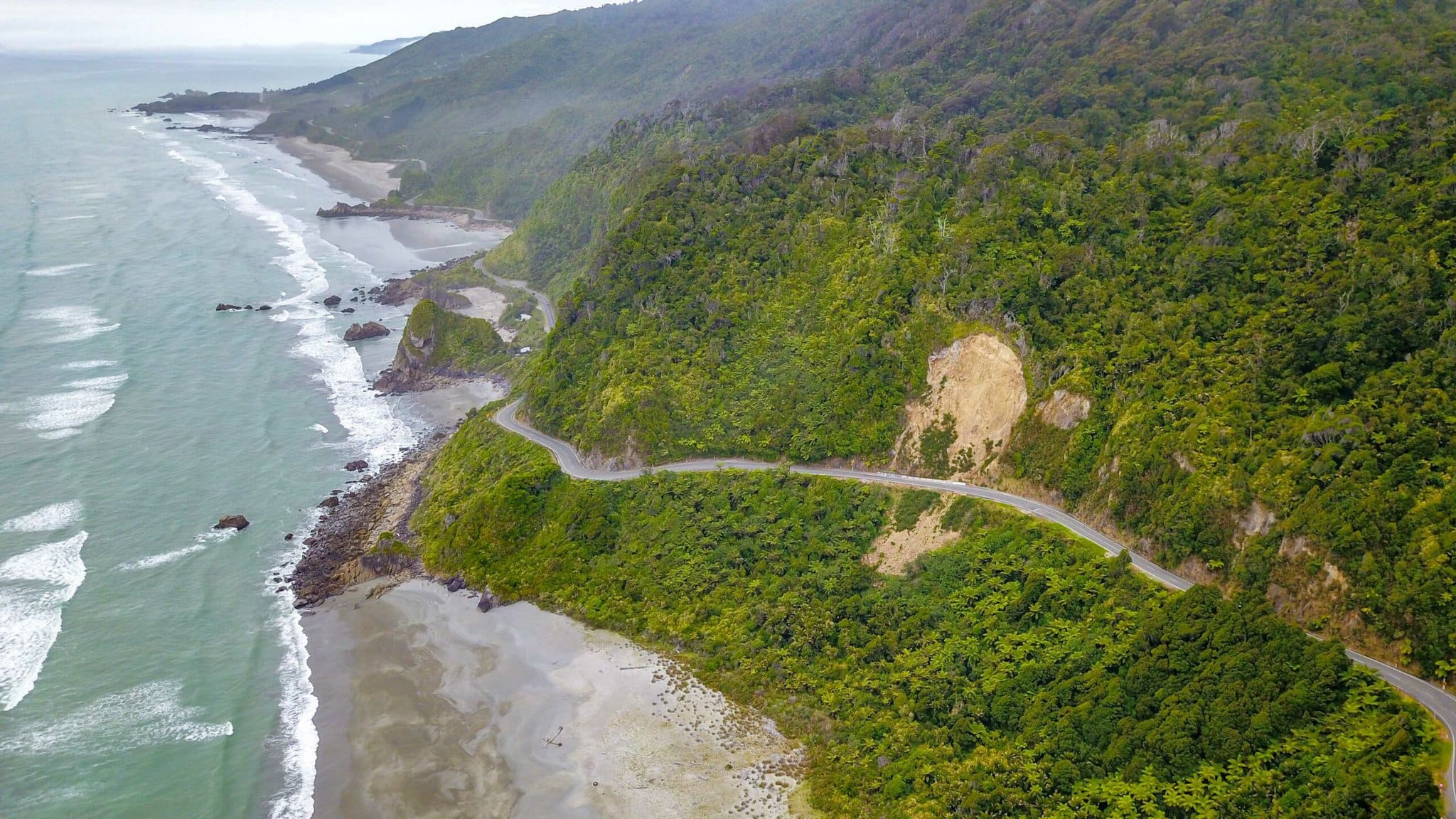 An aerial photo of a New Zealand South Island coastal road. The road is built along a long stretch of beach.