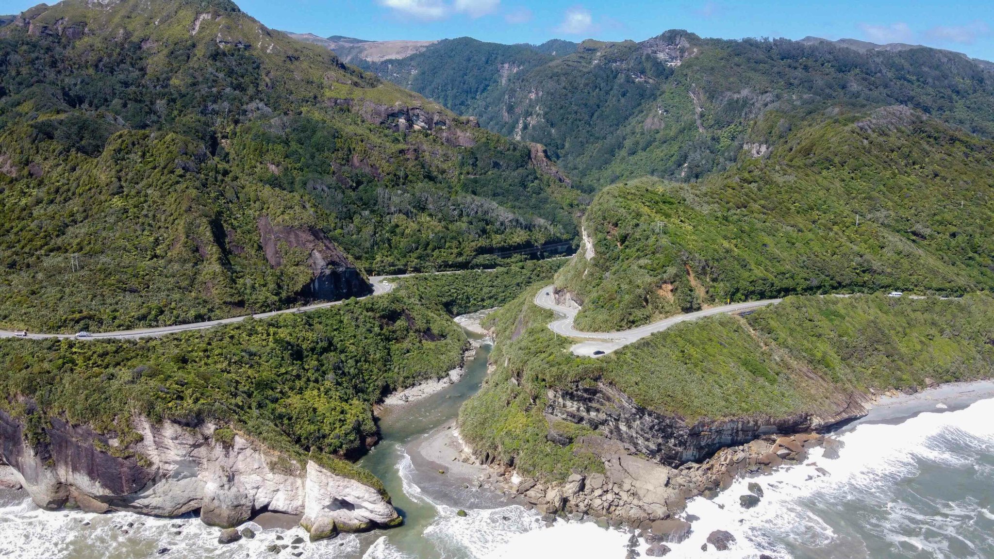 An aerial photo of a winding coastal road in New Zealand built into the side of steep hills beside the ocean.