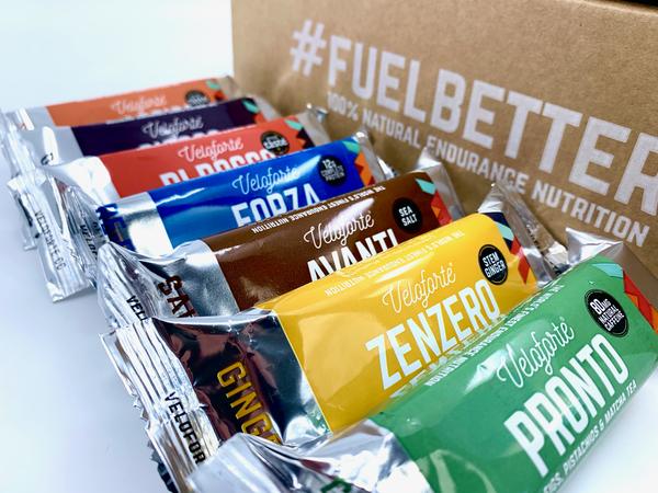 A variety of energy bars are laid out in a row in front of a Fuel Better sign.
