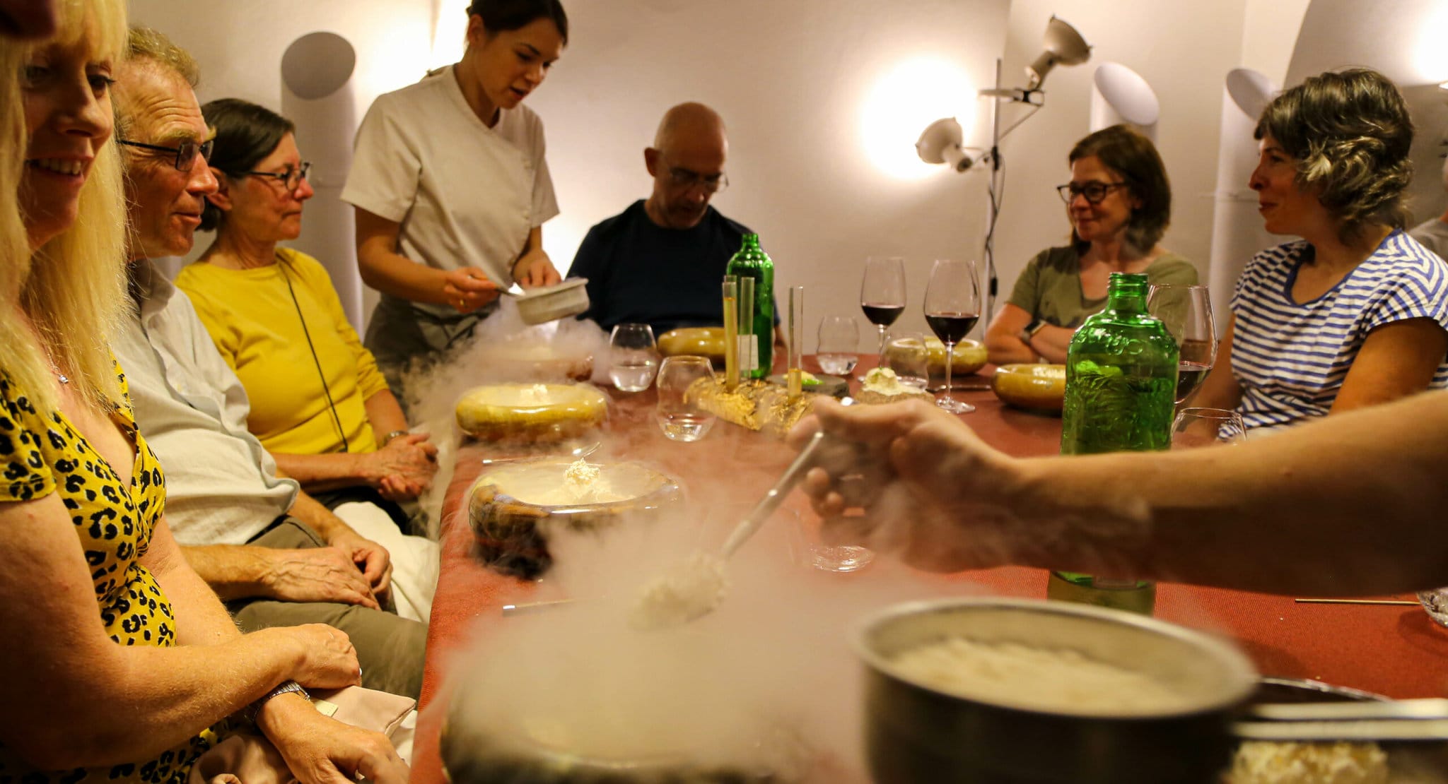 A group seated at a table laid with wine and plates. One person is being served by a waiter in a white uniform.