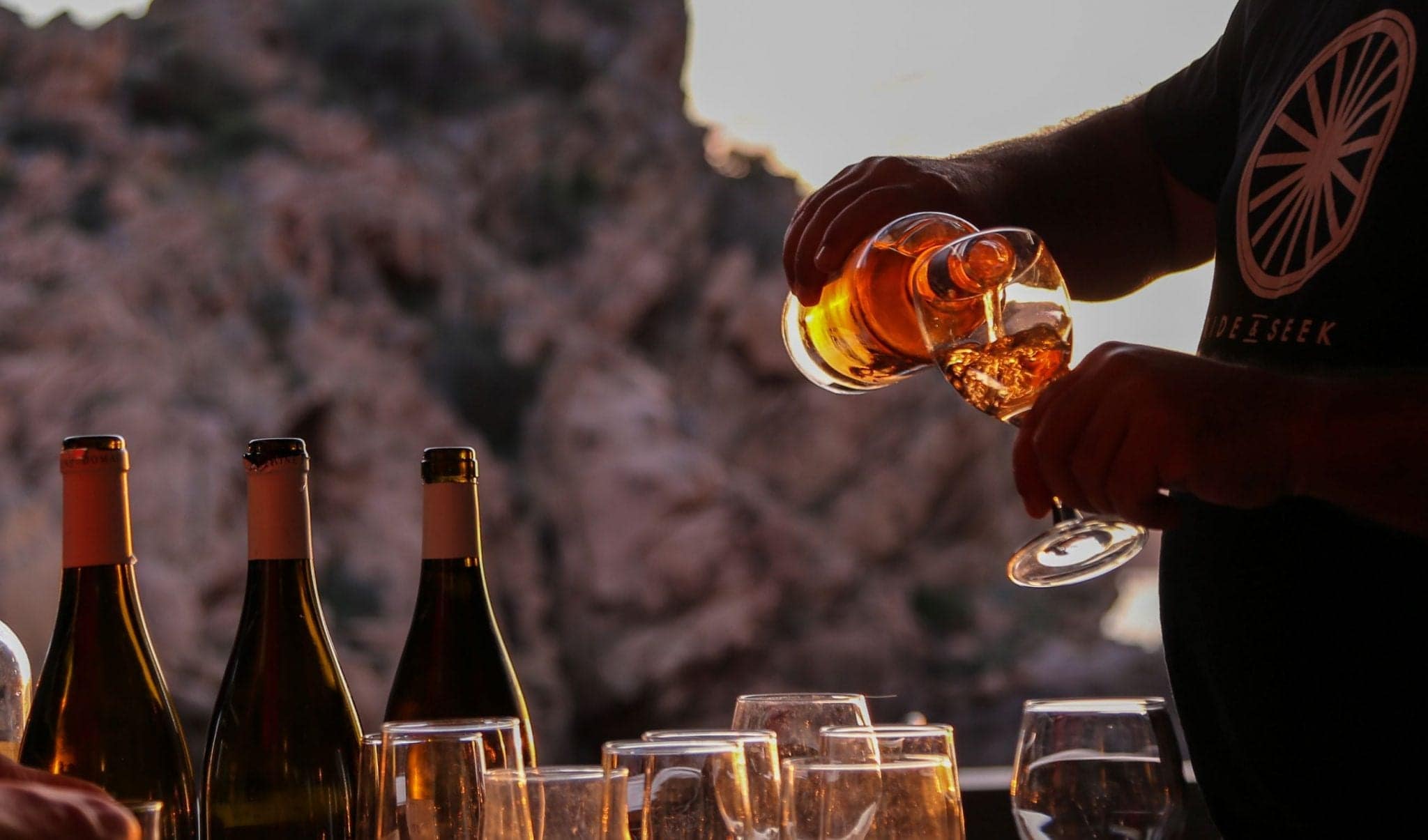 A partially visible person is pouring a glass of wine from a bottle. Other glasses and bottles are partially visible, with a large rocky cliff in the background.
