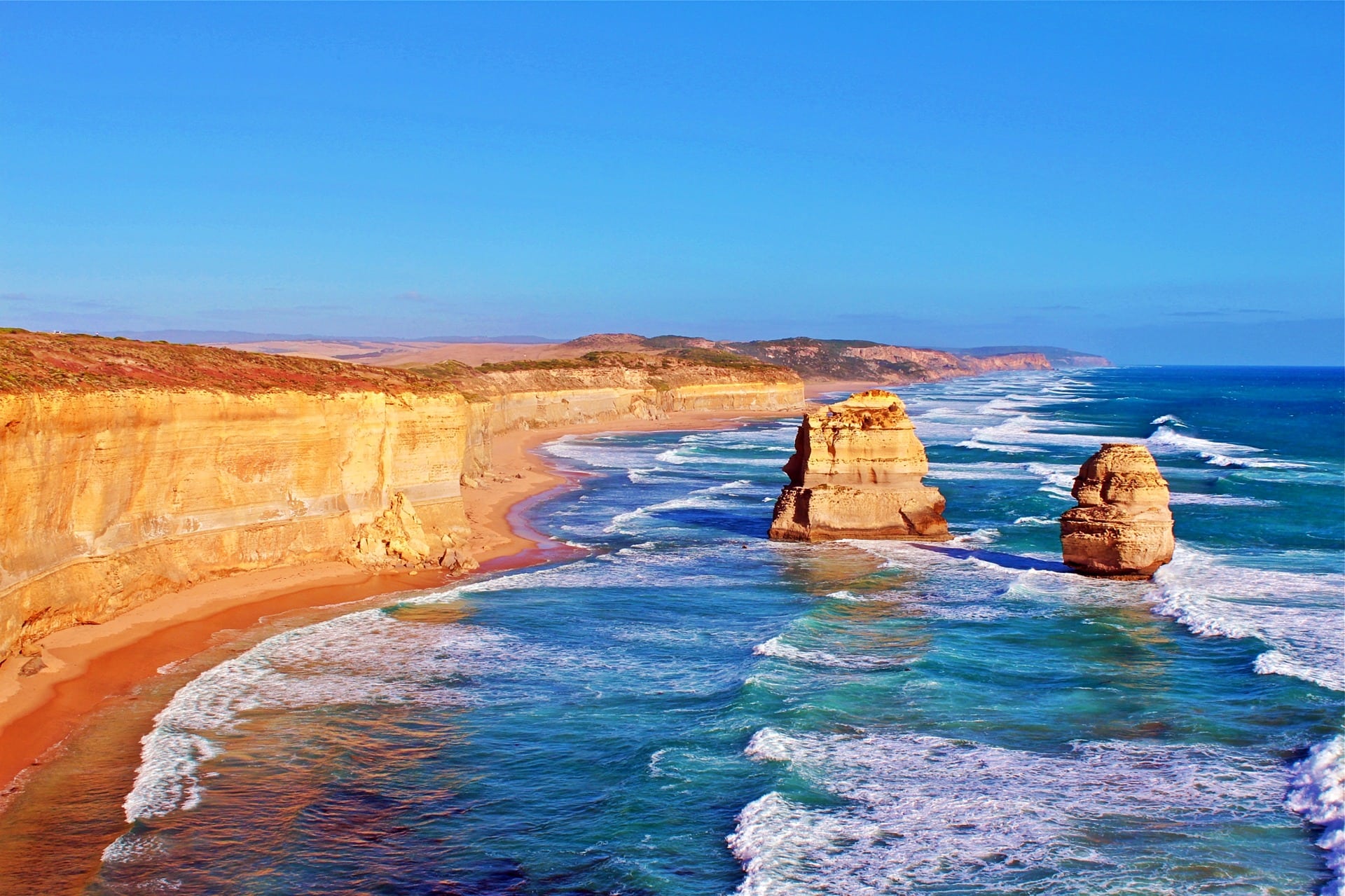 Two of the Twelve Apostles satnd in gently breaking waves with the staggering coastline stretching away into the distance.
