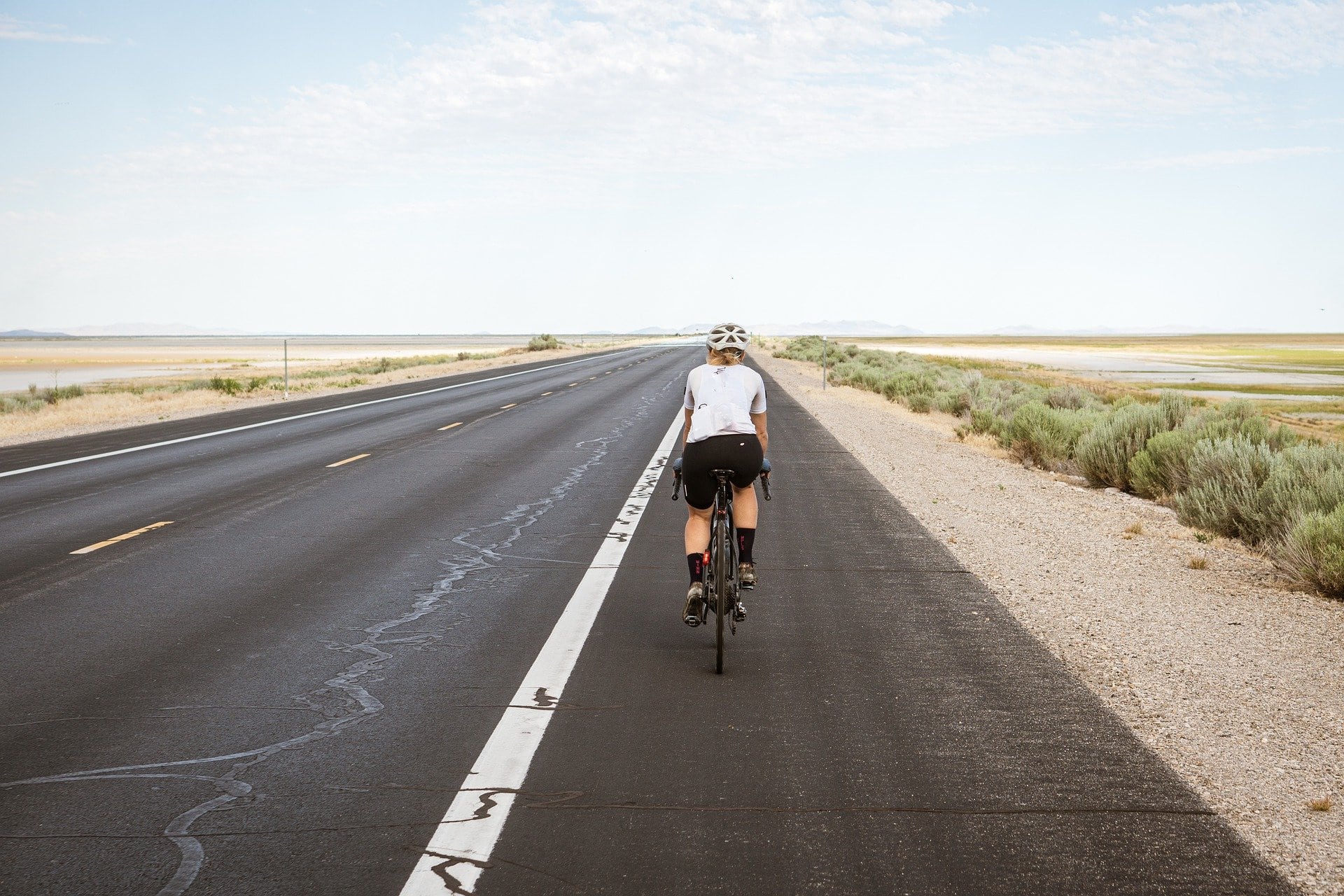 A cyclist rides away from the camera on the shoulder of a single-lane road. The road disappears away to the horizon in a dead flat, treeless landscape. Low shrubs line the right side of the road along the gravel edge.