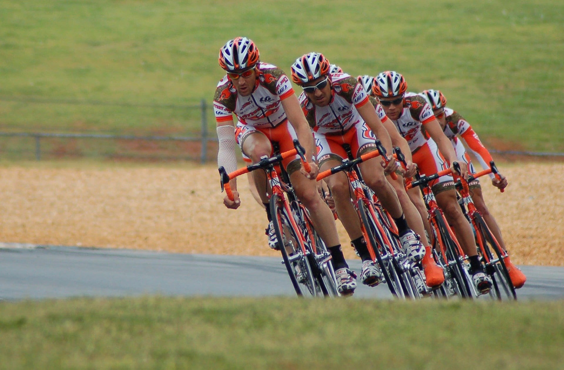 A team of cyclists rounds a corner on a wide curve on the road. A wide expanse of green grass fills the background.