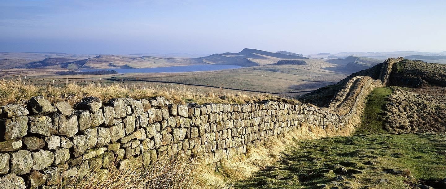 An old stone Roman wall stretches off into the distance following the lay of the green pastured hills. A blue lake surrounded by hills can be seen in the distance.