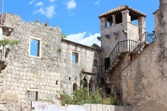 A square tower is perched on top of the ruins of a castle wall. Stairs with a metal stair rail lead up to the tower. An unkempt garden is in front of the bottom of the wall.