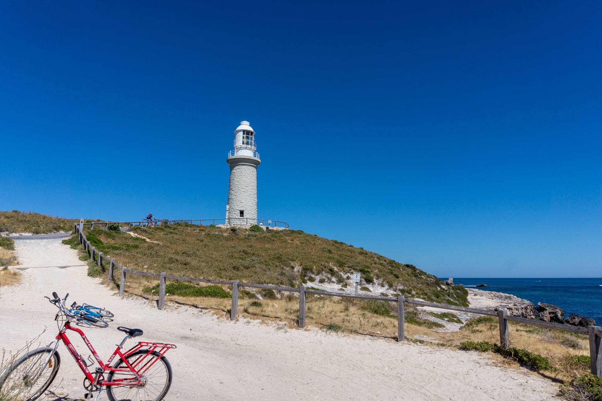 A white lighthouse sits atop a small hill overlooking the ocean. A red bike in the foreground is parked on a path leading off to the lighthouse.