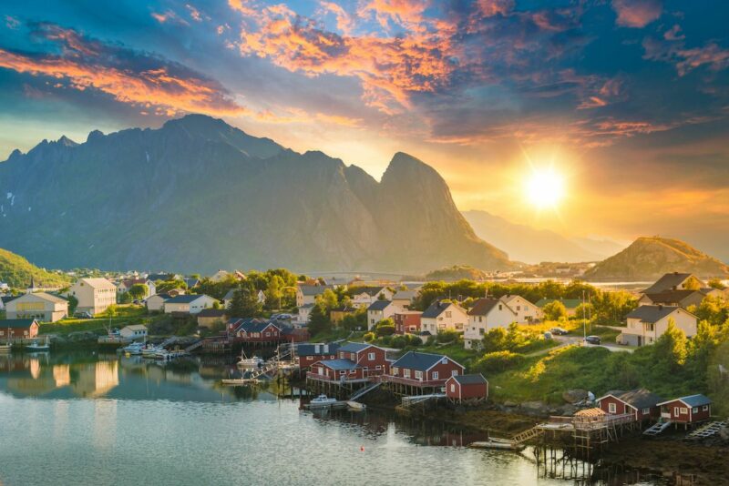 A small coastal village with red cottages that are built on stilts over the rocky edge of the water. A tall rocky outcrop is partially covered by grass in the background. The midday sun can be seen above the horizon.