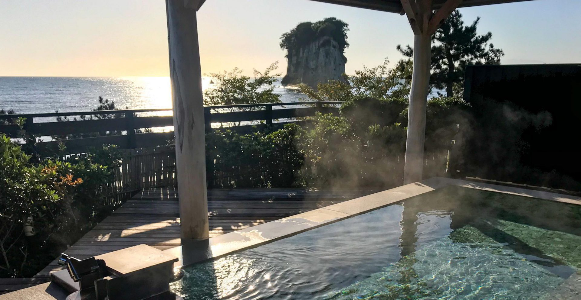 The photo looks over a steaming pool into the background of ocean stretching away to the horizon. The sun is low and reflecting off the water. A tall rocky outcrop stands out of the water.