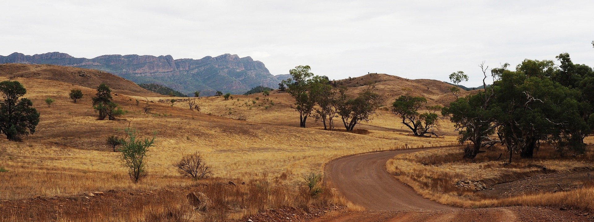 A gravel road winds through the dry, undulating, lightly wooded foothills in the foreground. An impressive, large rocky outcrop is in the far background.