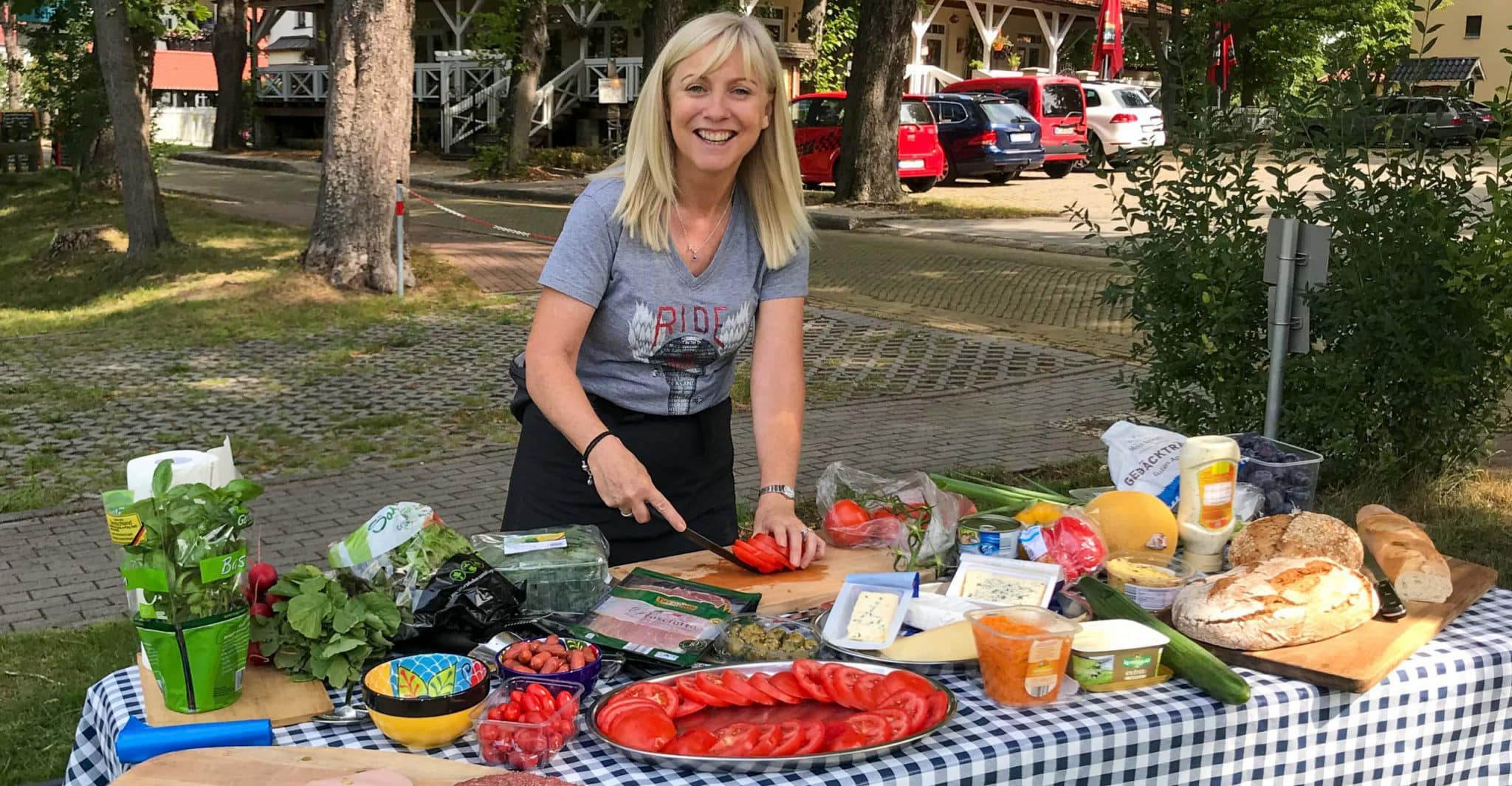 A person stands leaning over a picnic table laid with picnic food ready to be prepared. The person is holding a knife as if cutting a tomato already been sliced. The table is covered with a blue-checked tablecloth and is set up just off a quiet village street.
