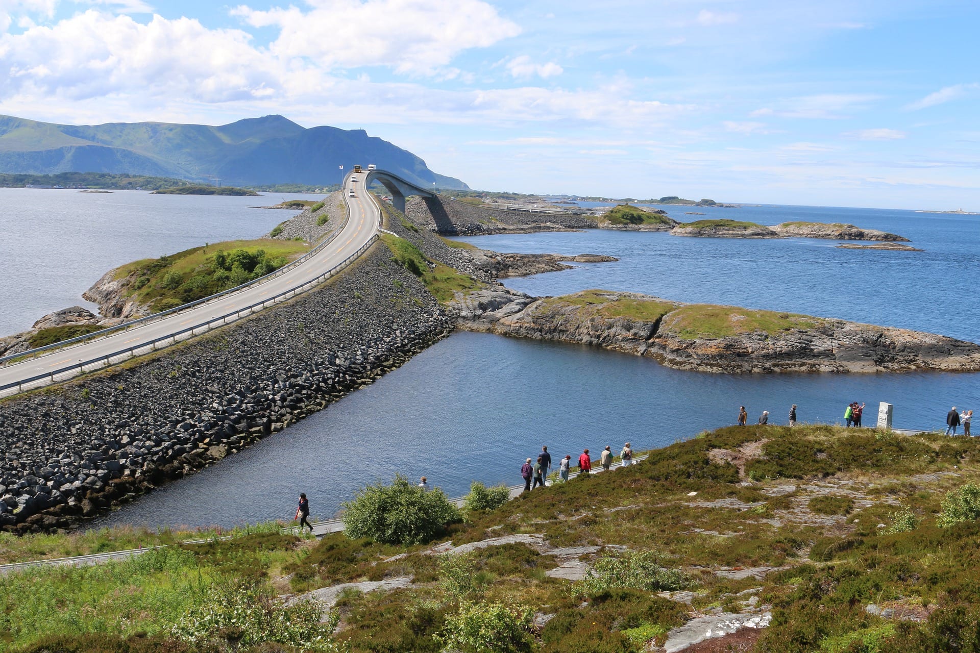 A built-up coastal road leads up to a bridge surrounded by water on either side. A large seaside hill rises in the background.
