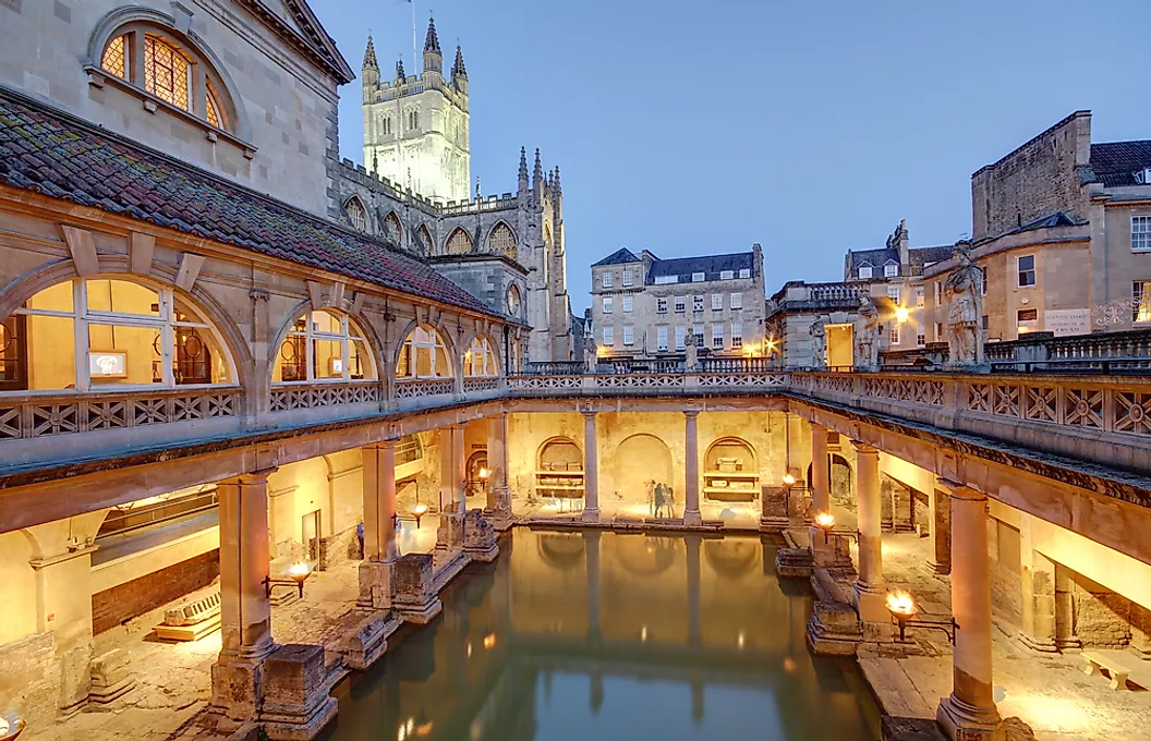 The buildings of the historical Roman baths surround a calm pool of water and are illuminated by soft lighting. The night sky is a pale blue in the background.