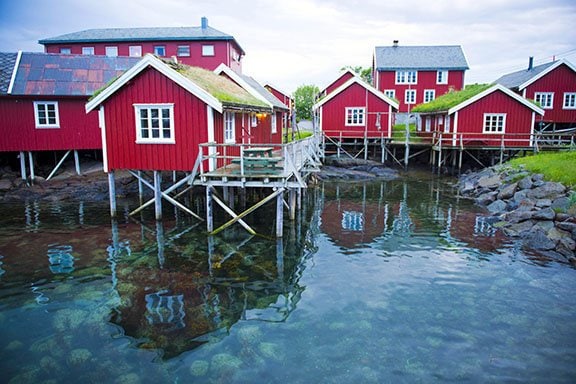 Several red cabins all with a light roof and window frames, built on stilts over the water.