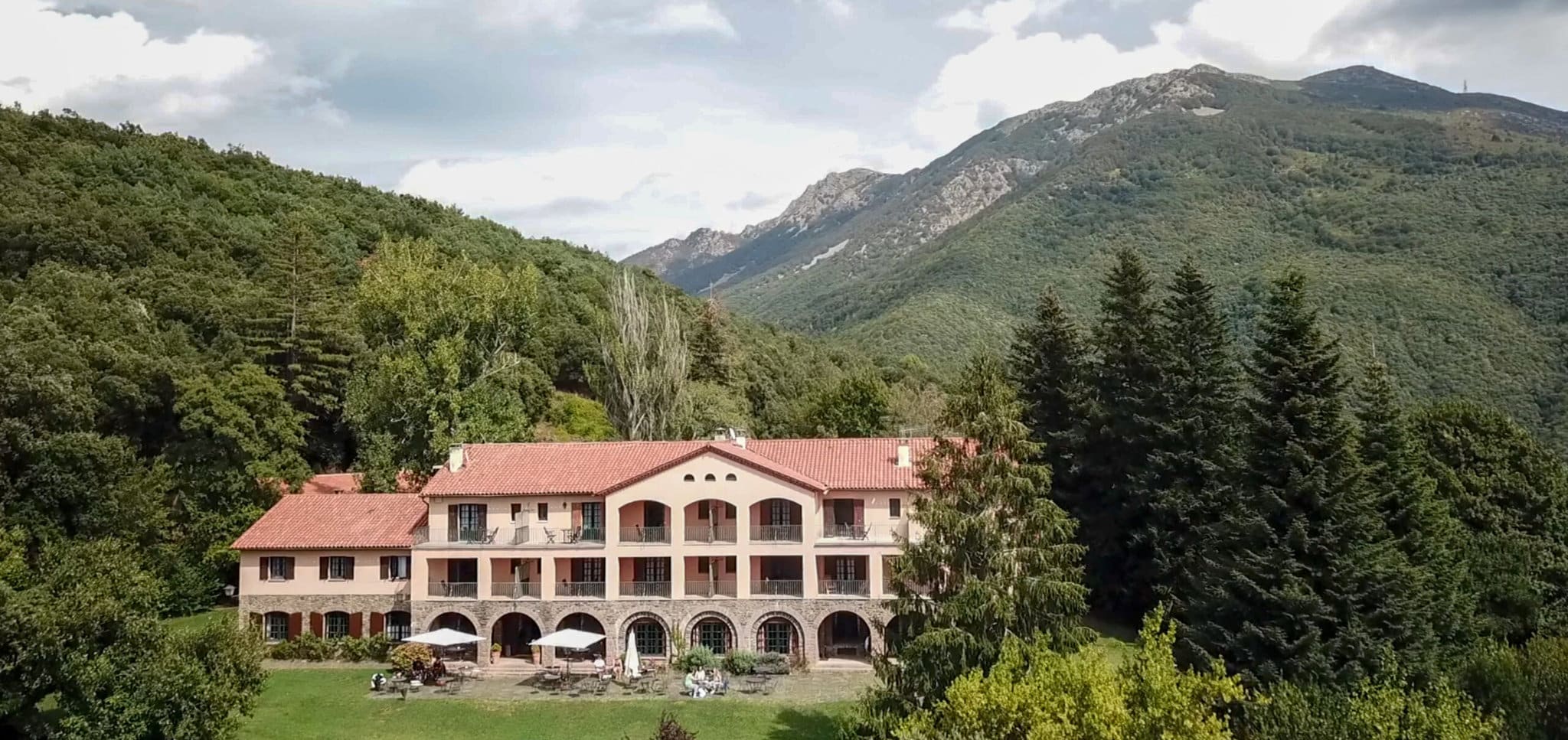 A red terracotta-roofed, multistory lodge is nestled in the hills, on a green lawn surrounded by forest with a tall hill in the background.