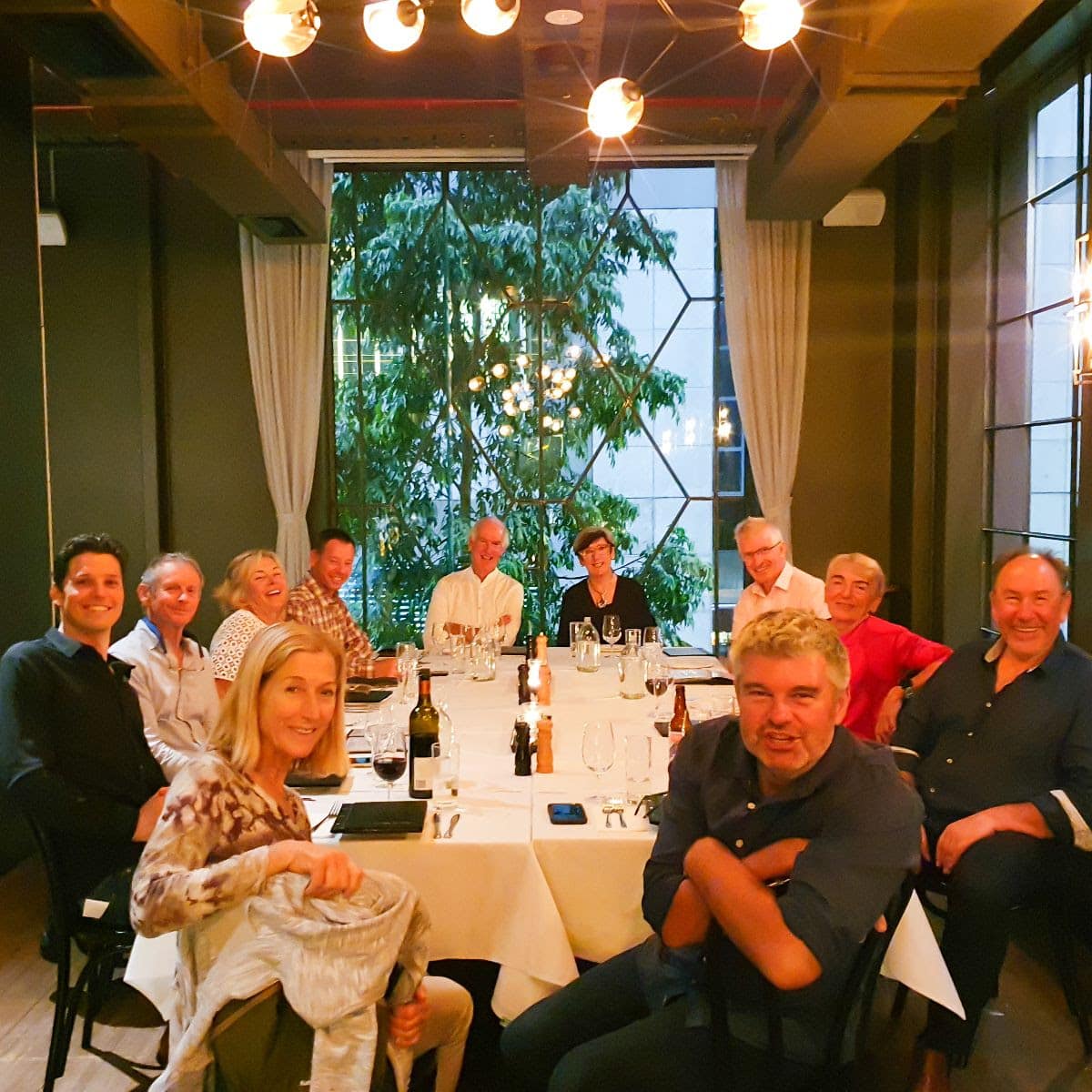 A group of Ride and Seek bike tour guests seated around a long rectangle table set with a white tablecloth. Lighting overhead a a picture window and gum trees in the background.