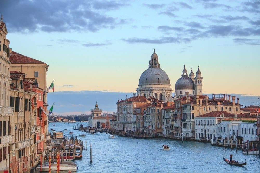A canal in the city of Venice with boats parked along the edge and gondolas and boats on the water. Two large domes rise above the old town buildings along the water's edge. A dimly lit sky with some cloud cover in the background.