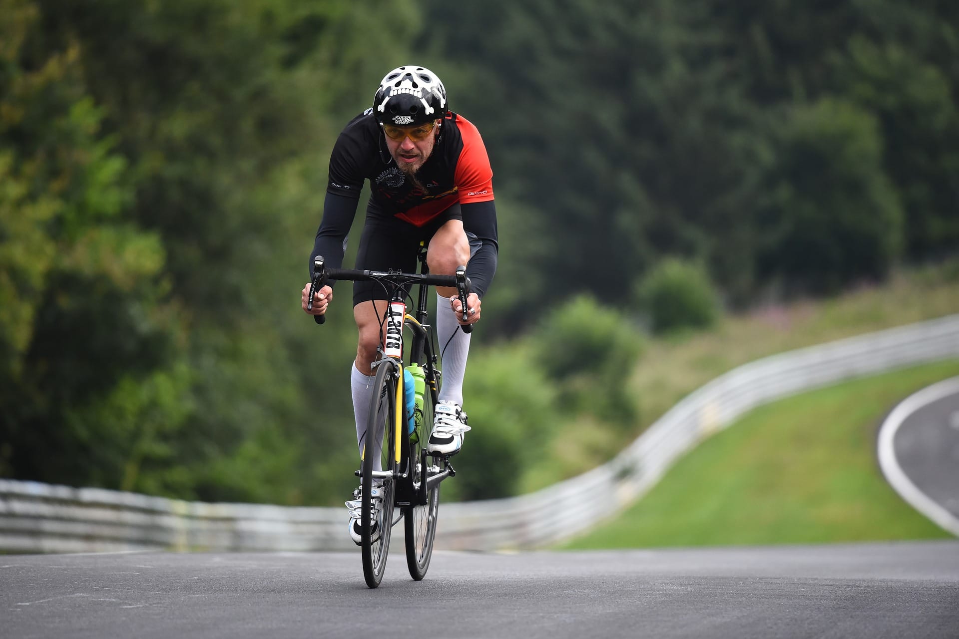 A cyclist rides on a racetrack towards the camera. A heavily wooded area fills the background.