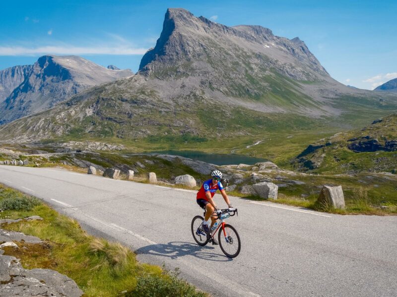 A lone rider cycles along the road as a stark mountain landscape rises in the background