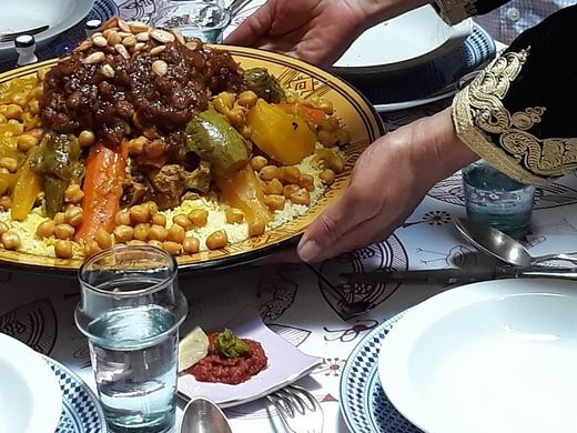 A large Moroccan couscous dish served with chickpeas, carrots, zucchini, peppers, and lamb, topped with caramelized onions and almonds, presented on a decorated platter.