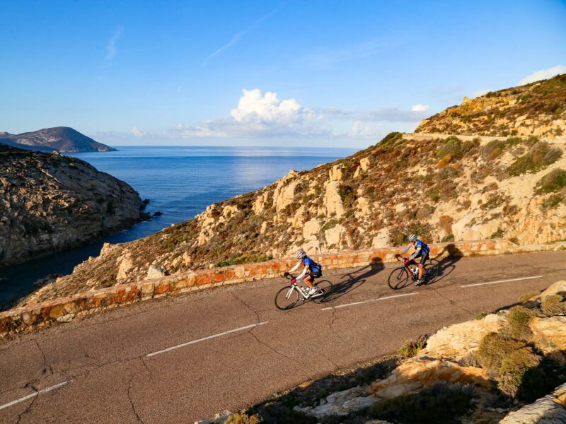 2 cyclists riding on a cliff edge road along the coast of Corsica overlooking the ocean