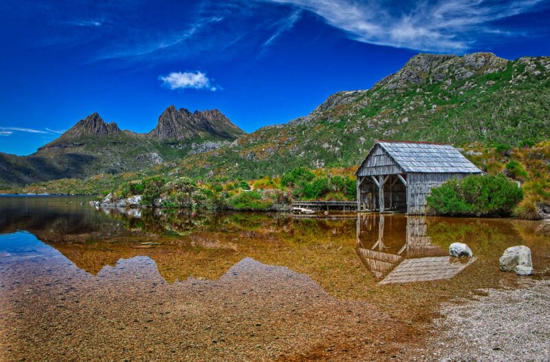 A wooden boathouse sits on the rocky shore of Dove Lake, with the rugged Cradle Mountain and cloudy sky in the background in Tasmania. The water reflecting the scene above it.