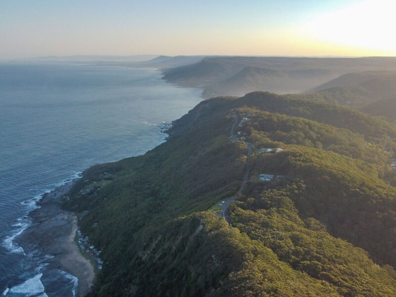 An aerial view of the Pacific coastline with a winding road on the side of a mountain ridge covered in a dense green forest, with waves from the ocean crashing on the rocky shore below.