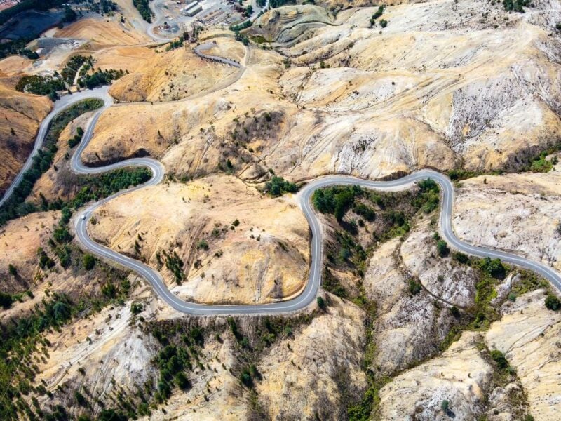 An aerial view of a winding road cutting through a barren, hilly landscape of orange, brown, and white earth, with small patches of green trees along the road at Queenstown, Tasmania.
