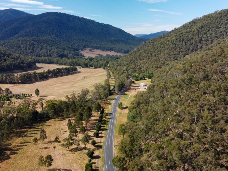 An aerial view of a straight road through a wide valley. The road is bordered by a line of trees on the left and a dense forest on the right, with a field in the foreground and a house in the distance. Large, forested mountains form the background.