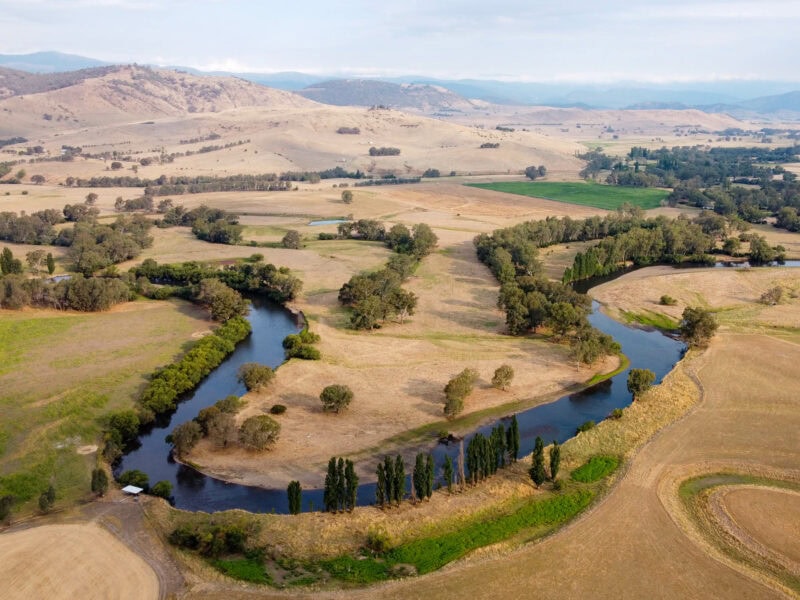 An aerial shot of a winding river in a wide, dry valley. The river is dark blue and curves through green and yellow fields, with scattered green trees and brown, grassy hills in the background.