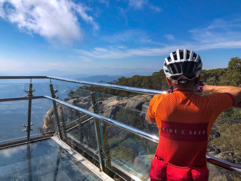 A person wearing a cycling helmet and an orange RIDE & SEEK jersey stands on a glass scenic lookout platform, looking out at a panoramic view of rolling mountains and a valley below under a blue sky.