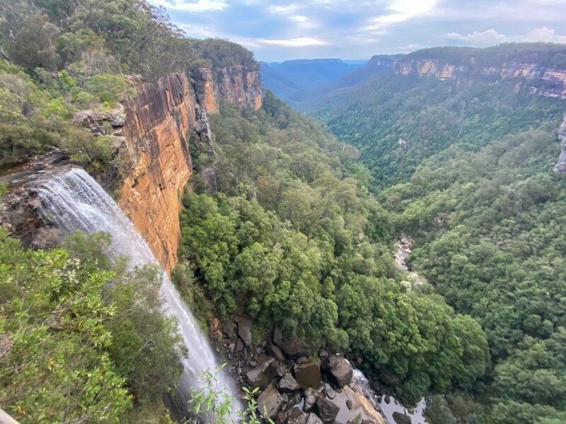 A scenic view of a tall waterfall cascading down a sheer cliff face into a deep, forested valley. The valley is filled with lush green trees and surrounded by other cliffs and mountains in the distance