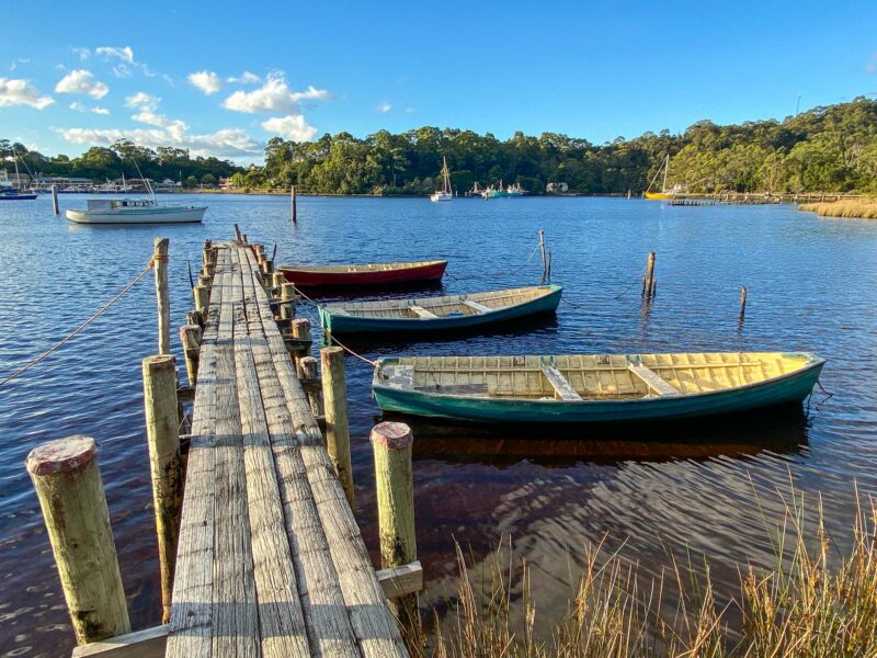 A low-angle shot of a wooden jetty extending into a calm body of water, with three small rowboats tied to it. The far shore is lined with lush green trees, and other boats are visible in the distance.