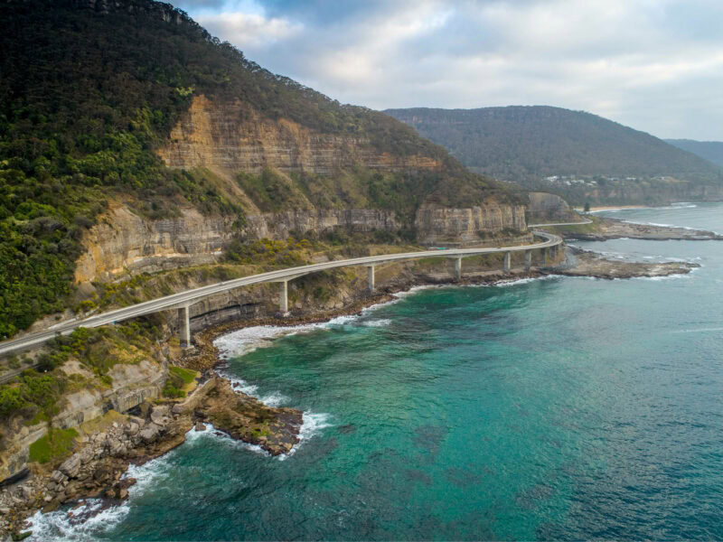 An aerial view of the Sea Cliff Bridge Wollongong, a curved road supported by white pillars that follows a steep, rocky cliff line above a turquoise ocean.