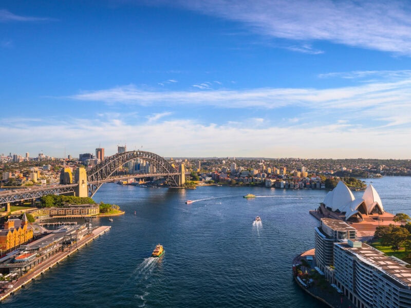 Daytime view of Sydney Harbour with the opera house and bridge on a partially cloudy day. There are ferries entering and leaving Circular Quay.