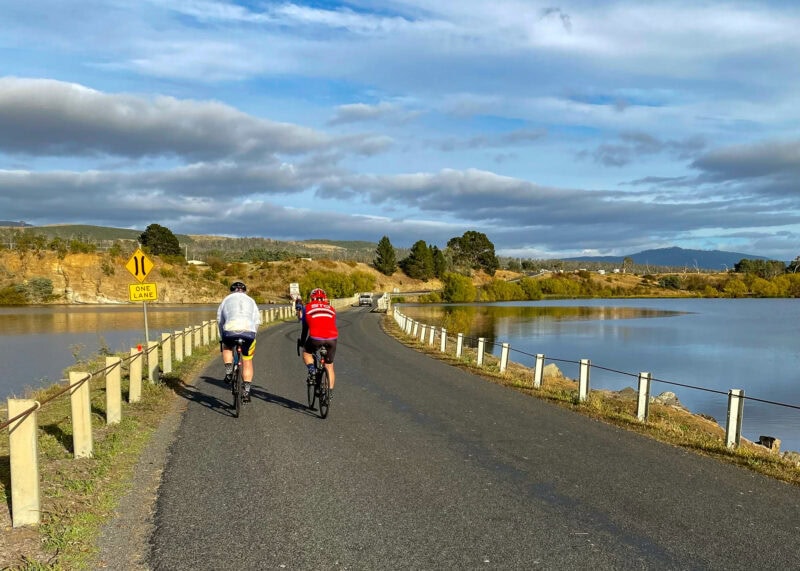 Two cyclists riding on a road with a lake on the right and a rocky embankment on the left, under a cloudy sky. A yellow sign on the side of the road reads "ONE LANE". There is a tall mountain in the background