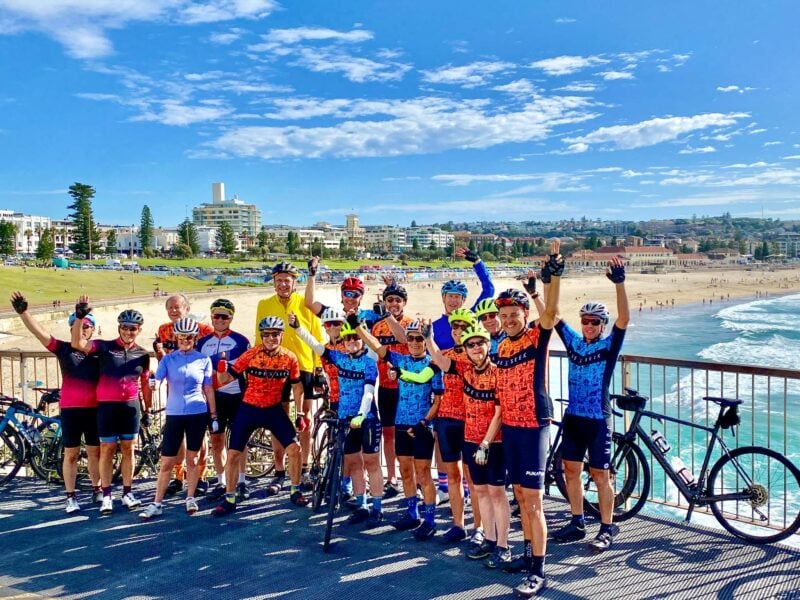 A group of cyclists pose with their bikes, hands raised high, with Bondi Beach stretching out into the distance in the back ground, on a clear blue sky day.