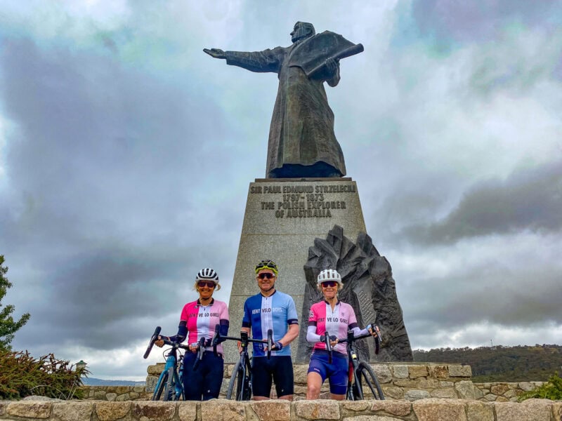 Three cyclists pose with their bikes in front of the Sir Paul Edmund Strzelecki statue in Jindabyne
