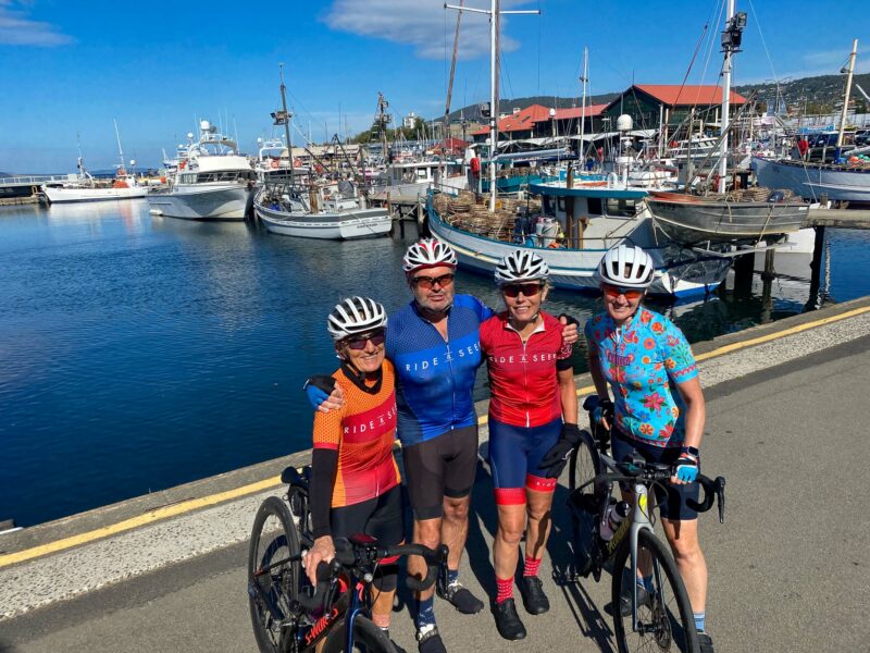 Cyclists pose for a group photo with their bikes in front of Constitution Dock, Tasmania. There are boats moored at the docks and some marine sheds in the background