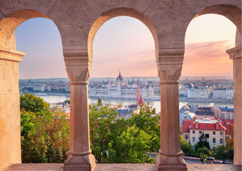 Photo looking through three arches if light coloured sandstone out to a city sitting behind a lake. There are bushes and a few houses in front of the lake.