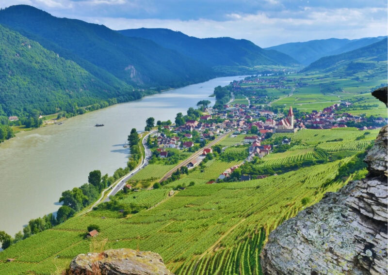 A village sits beside a wide river in a rural scene. On the opposite side of the river is a row of hills.