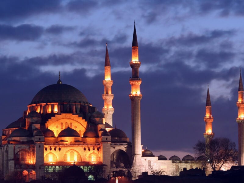 A mosque is lit up, with an evening sky behind.