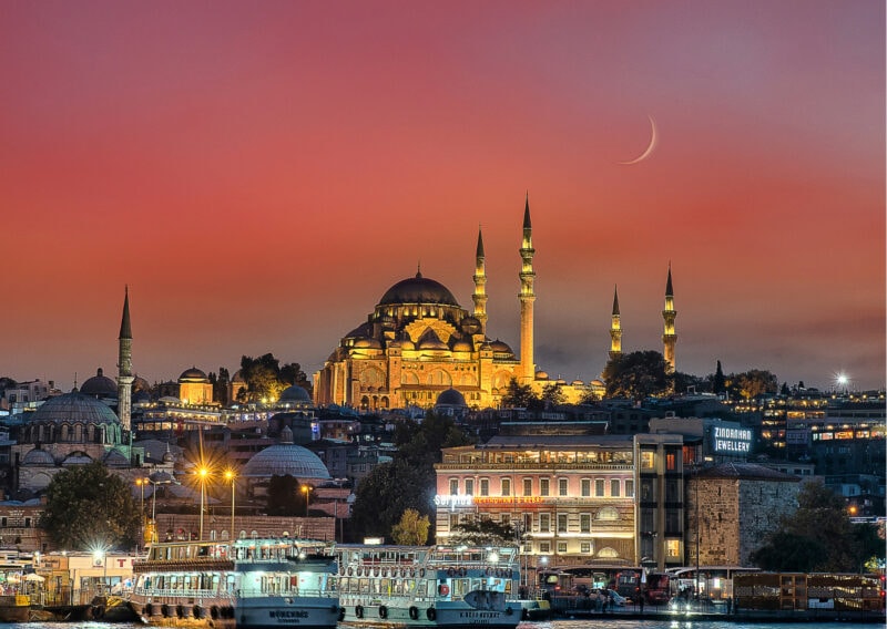 A mosque with four minarets is lit against the old town with a vivid sunset in the background.
