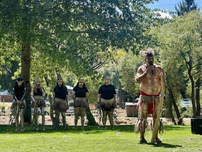 A man dressed in traditional a Bangerang Indigenous costume stands holding a microphone on a grassy patch. Behind him in the shade of a large tree is a group of Indigenous dancers also dressed in traditional costume.
