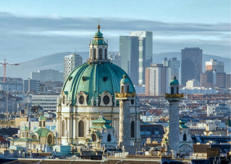 The blue dome of an old town cathedral with two minarets in front, sits amongst modern city buildings. A blue cloudy sky behind.