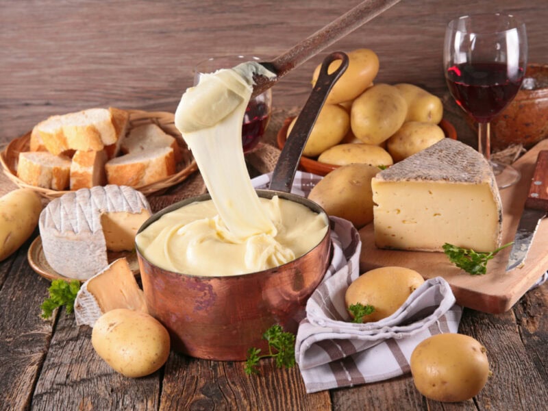A spoon that has been dipped in melting aligot which is now is oozing back into the pot below. Surrounding this is an array of breads.