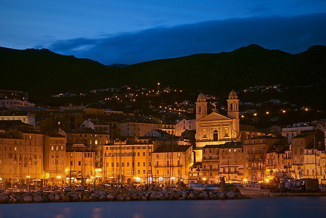 An evening scene of an old town set on the edge of the water. The buildings are illuminated and a dark, cloudy sky behind.