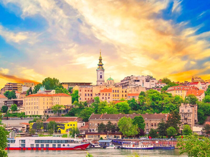 A golden sky behind an old city landscape set beside a river. River cruise boats float on the river. A tall steeple rises out of the old buildings.