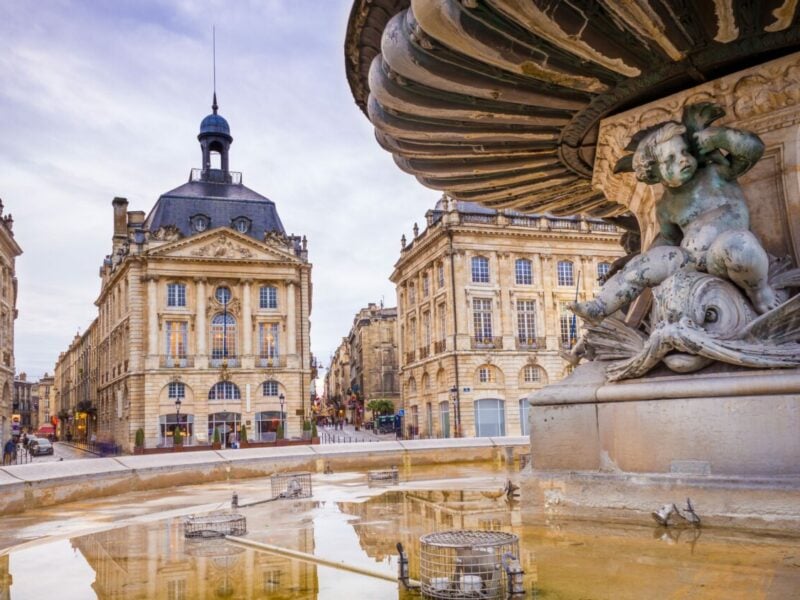 Two multi-story old town buildings are set behind an old town square fountain filled with water.